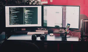 desk setup with two monitors, keyboard and a pair of headphones with a smartphone on a stand on the right-hand side