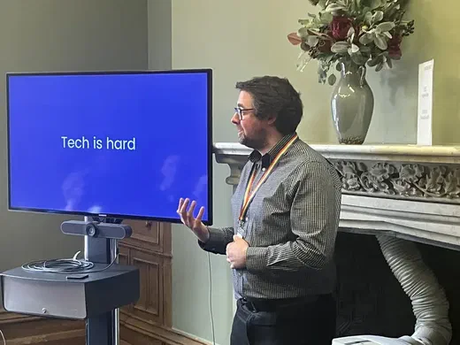 Richard Jackson presenting a slide reading 'Tech is hard' beside a fireplace with a flower arrangement.