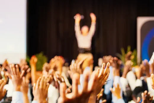 A speaker on stage with raised arms addresses a crowd with hands raised in the air.