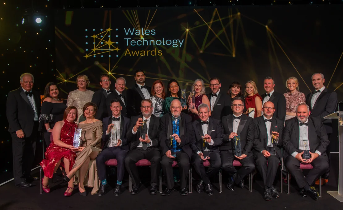 A large group of people in black-tie formal wear, sitting and standing in a group, holding their awards