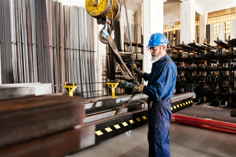 A production operative using an overhead crane to move steel bar in a metals warehouse