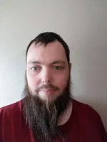 Mathew, a white man with short brown hair and a long brown beard, is looking toward the camera, in front of a white background