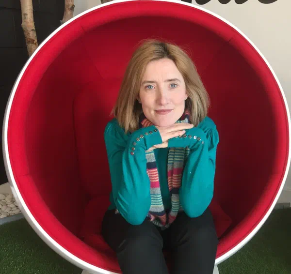 Ruth, a pale woman with shoulder-length strawberry-blonde hair, sitting in a red egg chair.