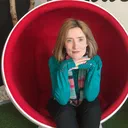 Ruth, a pale woman with shoulder-length strawberry-blonde hair, sitting in a red egg chair.