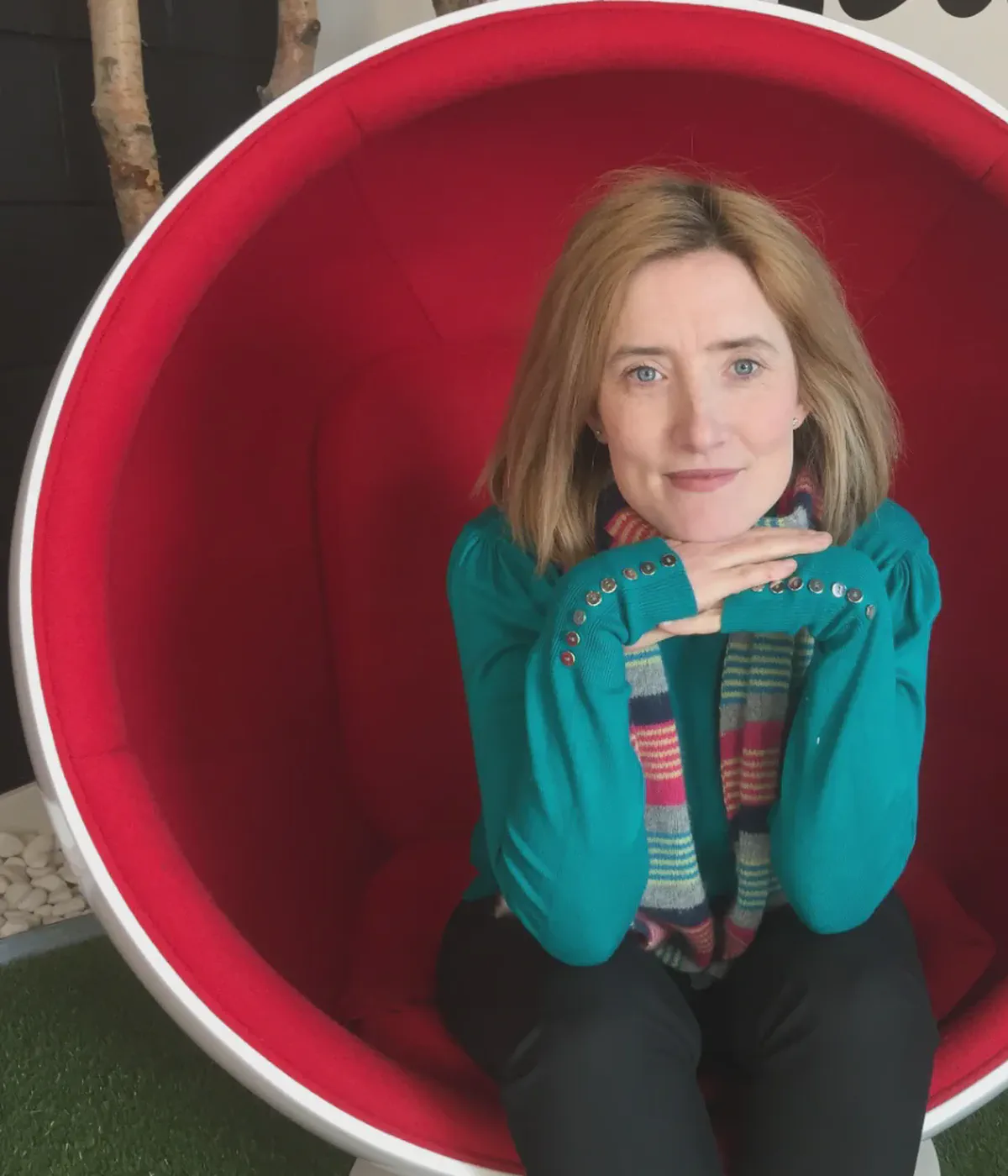 Ruth, a pale woman with shoulder-length strawberry-blonde hair, sitting in a red egg chair.