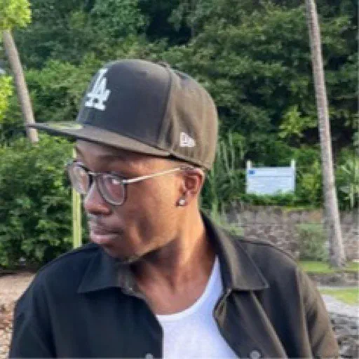 Tyree, a Black man with short cropped facial hair, black rimmed glasses, and a black LA cap, is facing to the side in front of some trees