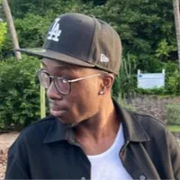 Tyree, a Black man with short cropped facial hair, black rimmed glasses, and a black LA cap, is facing to the side in front of some trees