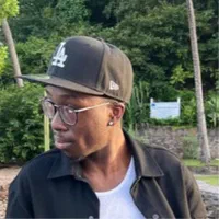 Tyree, a Black man with short cropped facial hair, black rimmed glasses, and a black LA cap, is facing to the side in front of some trees